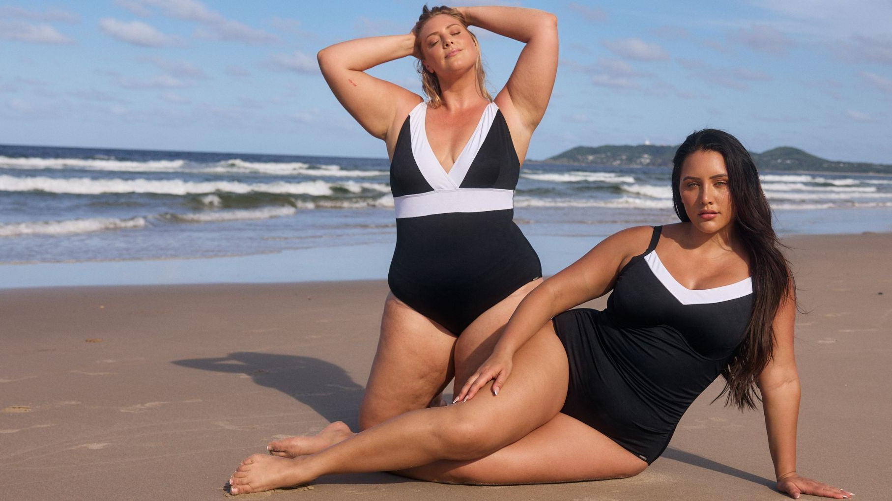 Two women pose on the beach wearing black and white one piece swimsuits