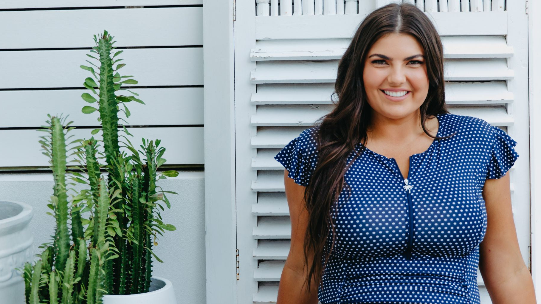 Woman with long dark brown hair wears navy and white polkadot swimsuit with zipup and frill sleeves