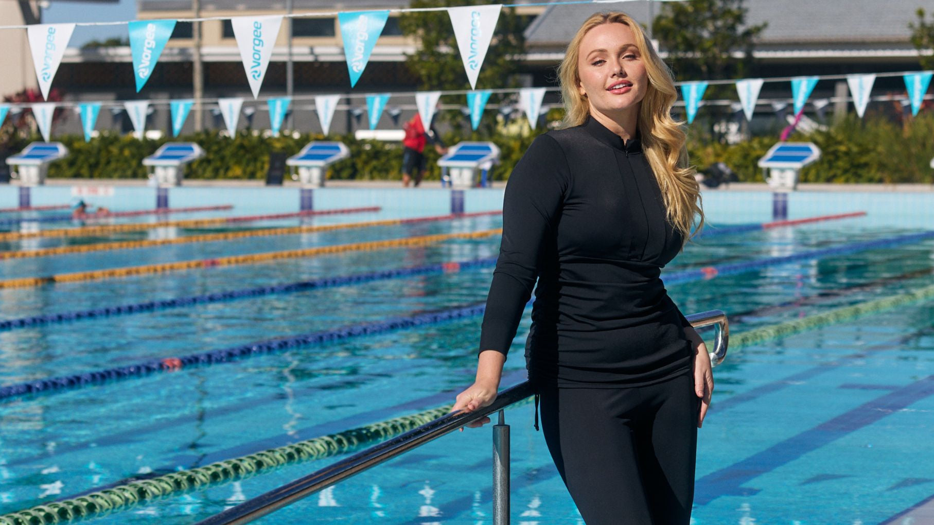 Woman with long blonde hair poses by the pool wearing black long sleeve rash vest with full length swim leggings.