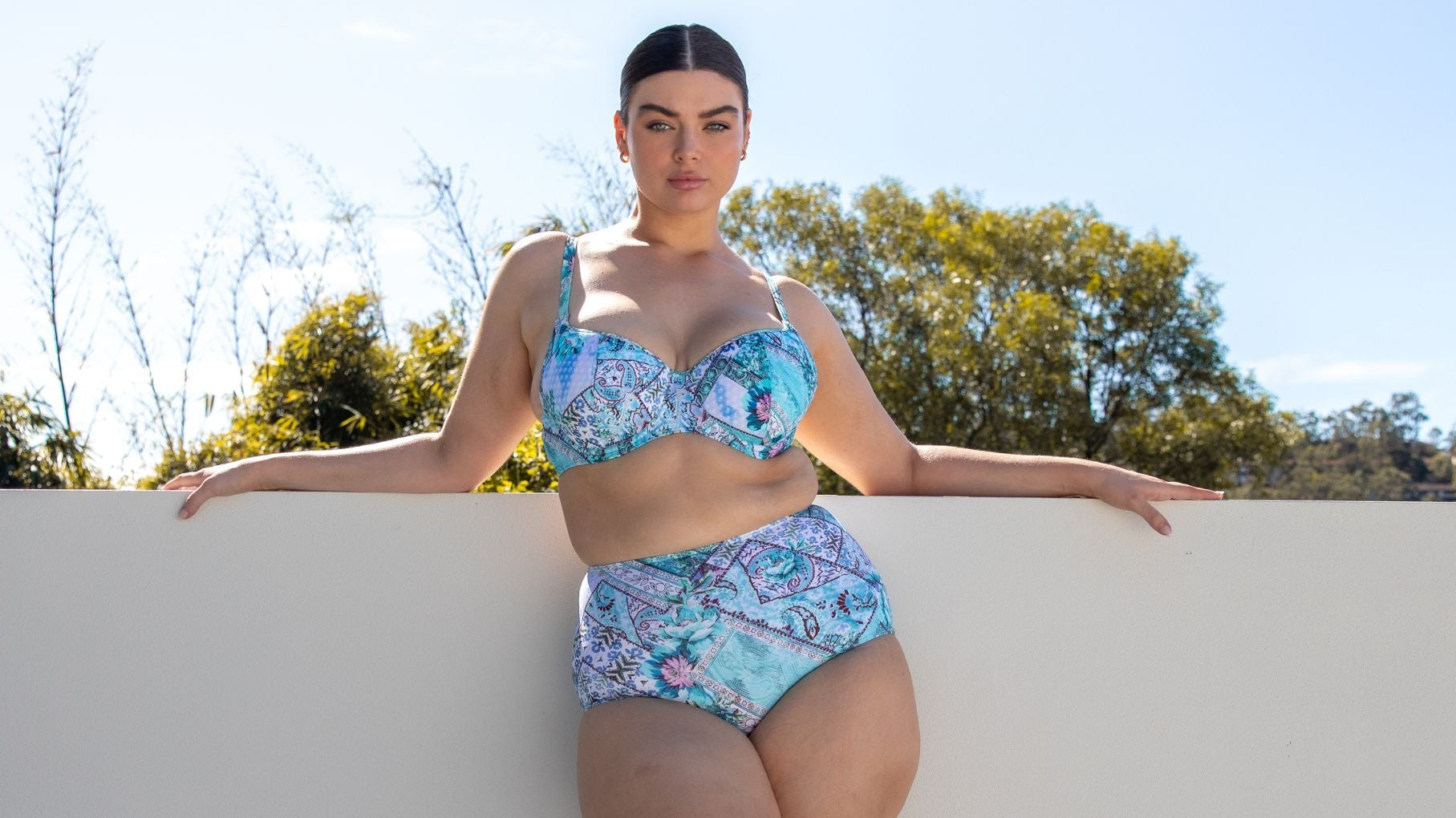 Woman with slicked back brown hair poses against a white wall wearing Whitehaven blue patchwork underwire bikini top and high waisted pant