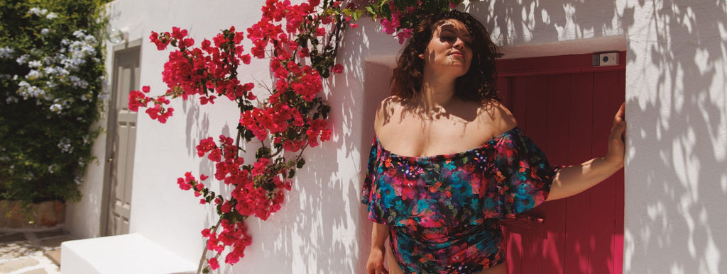 Brunette woman poses by a pink door under a canopy of bougainvillea wearing Midnight Garden off the shoulder one piece swimsuit