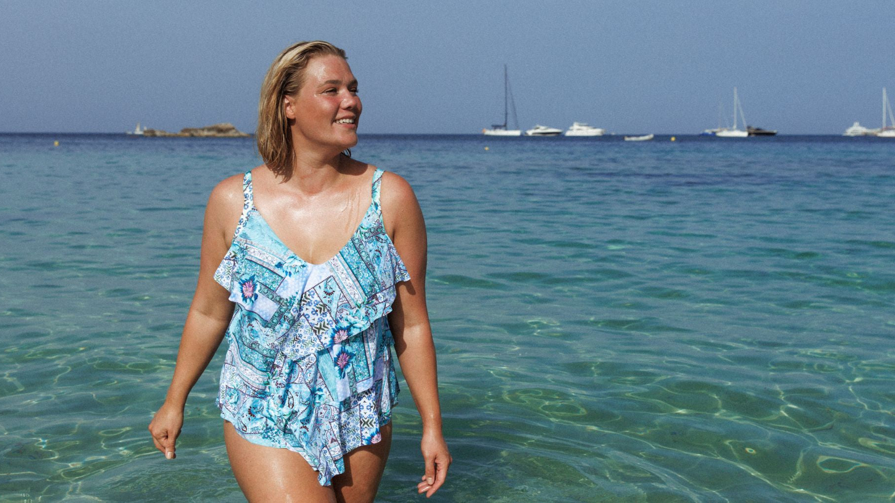 Woman with short blonde hair wades through the water wearing Whitehaven blue patchwork print 3 tier tankini top and matching pants.