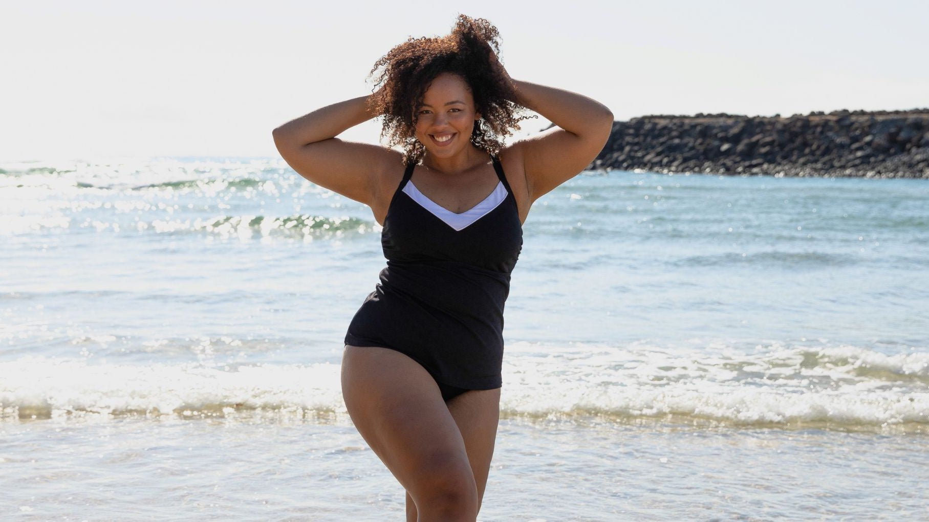 Woman with curly brown hair poses on the beach wearing black tankini top with white v neck trim