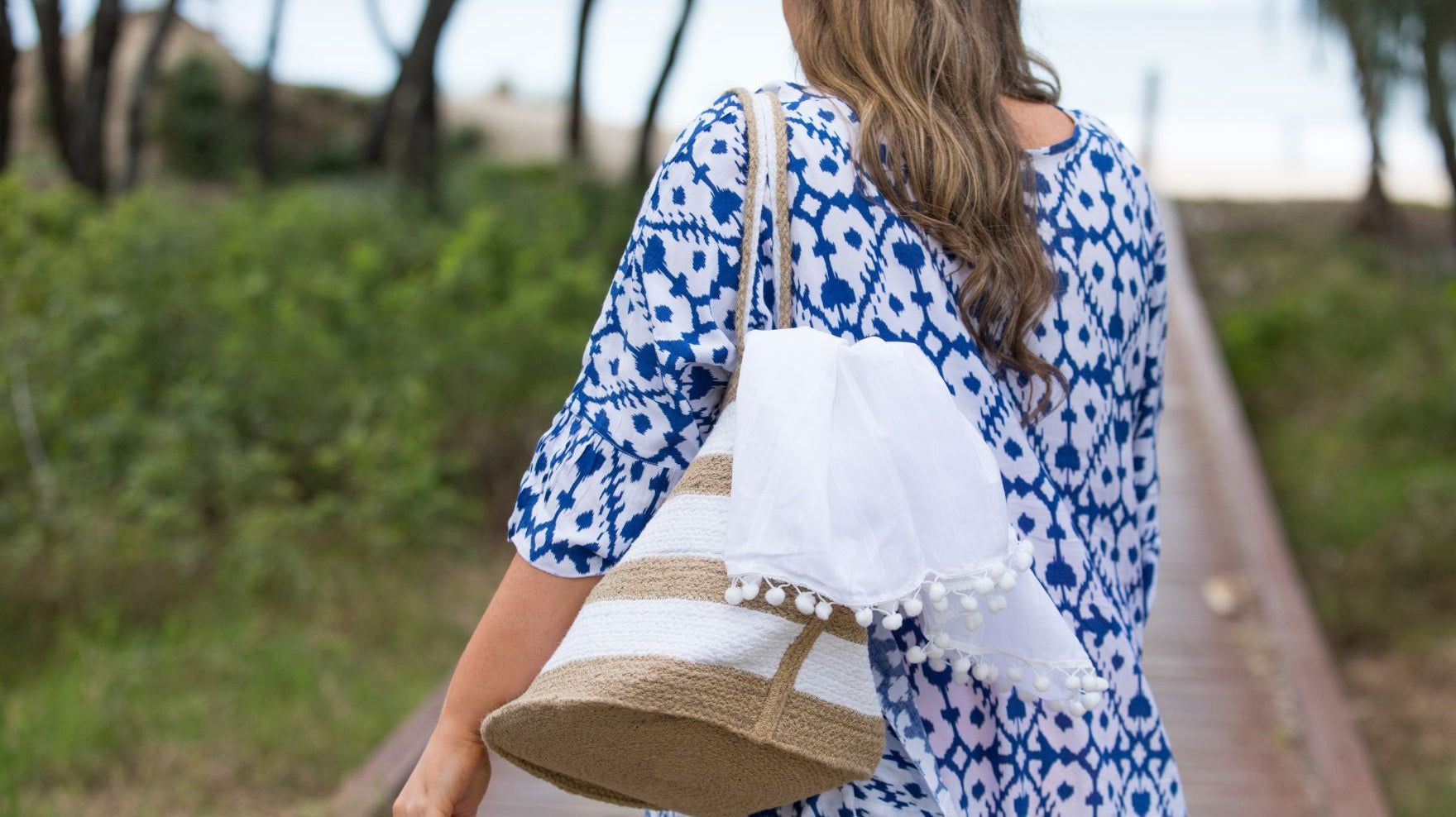 Woman wearing a blue and white patterned kaftan walks towards the beach carrying a natural and white striped jute beach bag