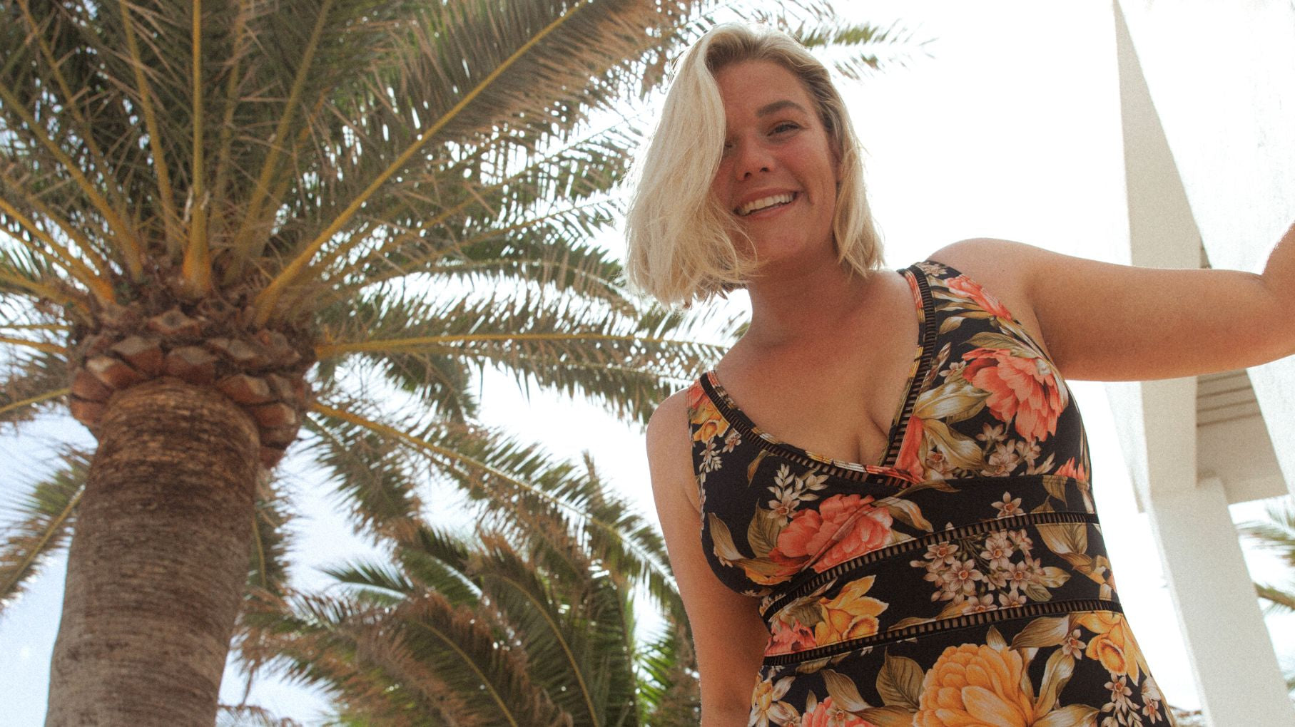 Woman with blonde hair poses by a palm tree wearing Frenchy Black floral V neck one piece swimsuit