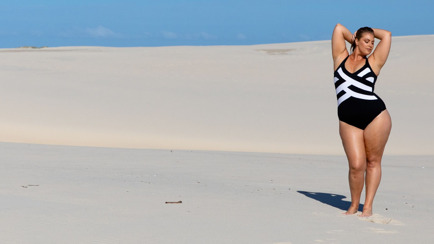 Woman poses on a sand dune wearing a black and white criss cross one piece swimsuit