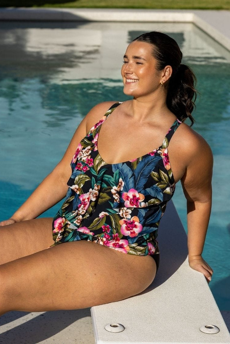 A woman wearing a floral Tiered tankini swimsuit sits on the edge of a swimming pool outdoors.