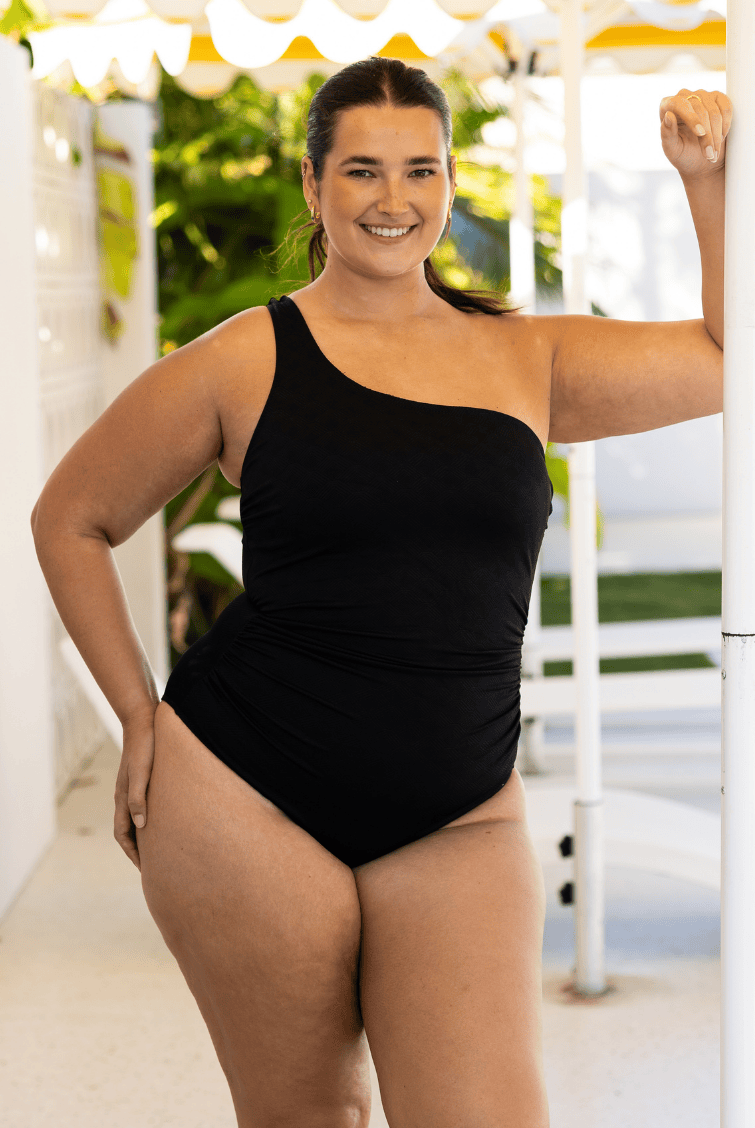 Woman wearing a black one-shoulder swimsuit standing outdoors.