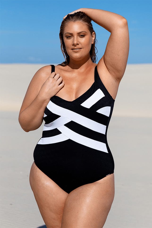 Plus size model stands on sand dunes in a black and white one piece swimsuit with criss cross pattern along the torso