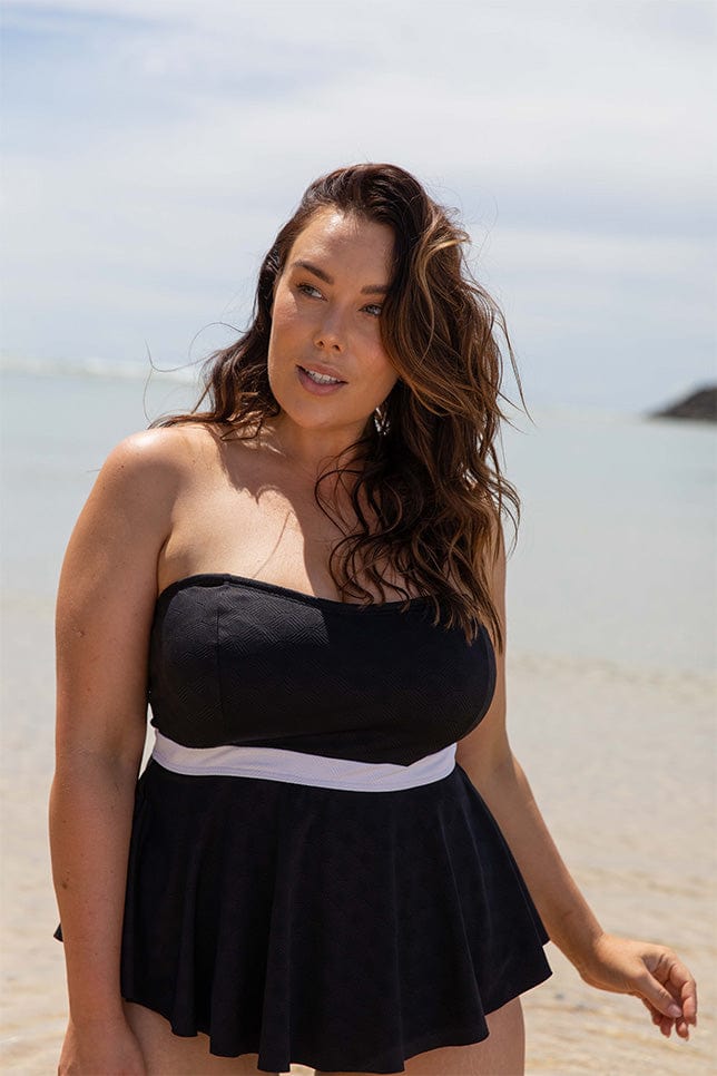 Brunette woman stands on the beach in a bandeau tankini top. The tankini has a white band along its waist and goes out like a peplum on the bottom. This swimsuit top can be worn with or without straps