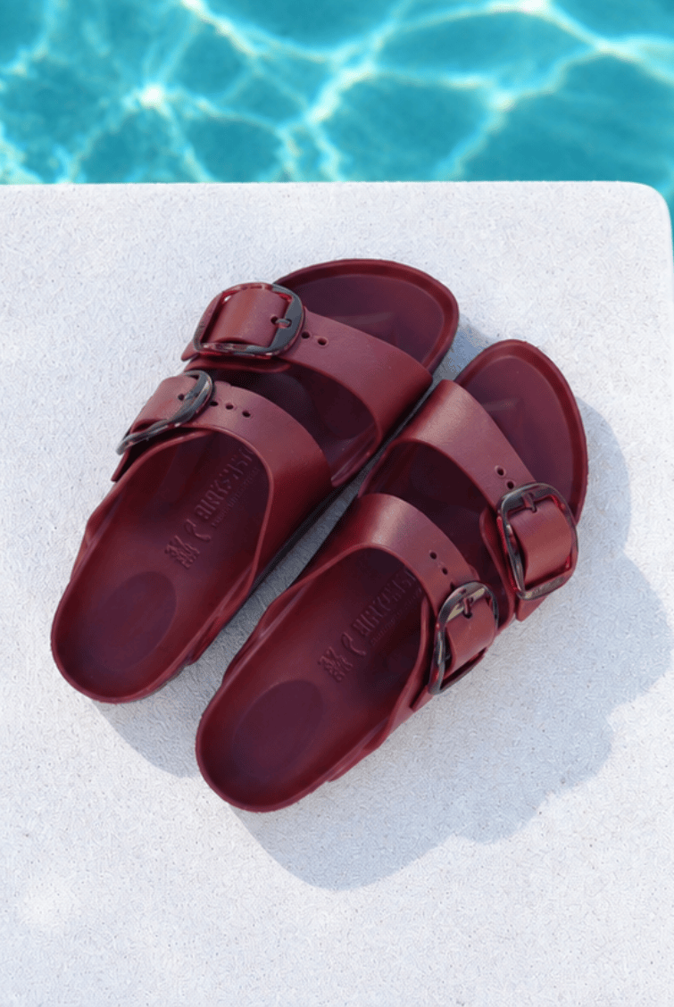 A pair of burgundy double-strap slide sandals with dark metal buckles, displayed on a white stone surface with a swimming pool background.