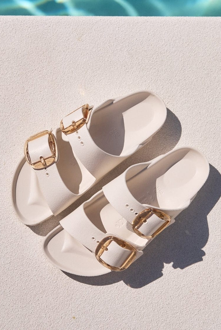 Pair of white sandals with gold buckles on a diving board over a pool.