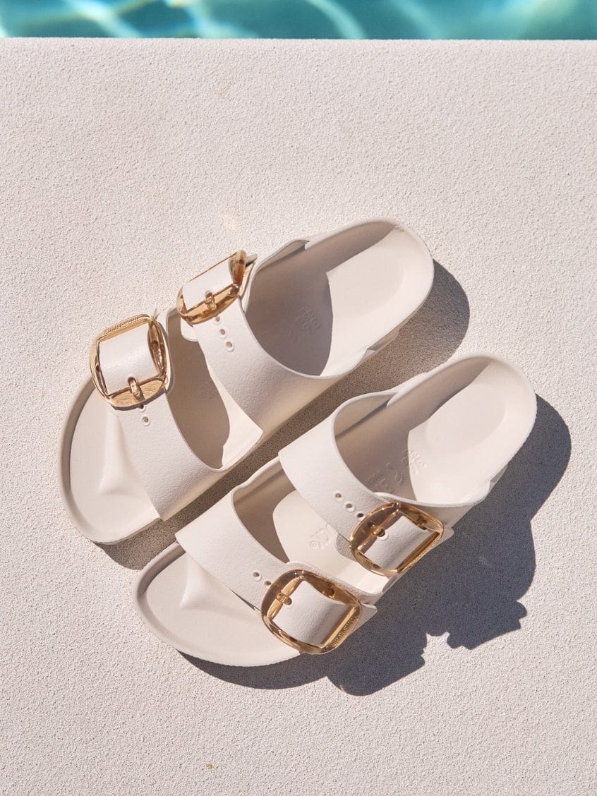 Pair of white sandals with gold buckles on a diving board over a pool.