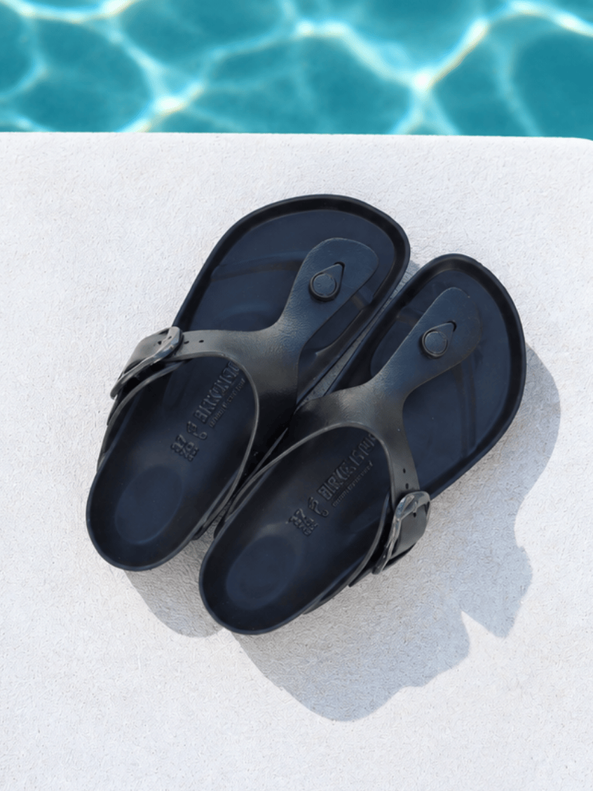 A pair of black rubber thong sandals with adjustable buckle straps, displayed on a light concrete surface with a swimming pool background.