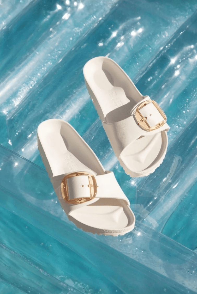 A pair of off-white slide sandals with gold-tone buckle details, arranged on a translucent blue inflatable pool float.