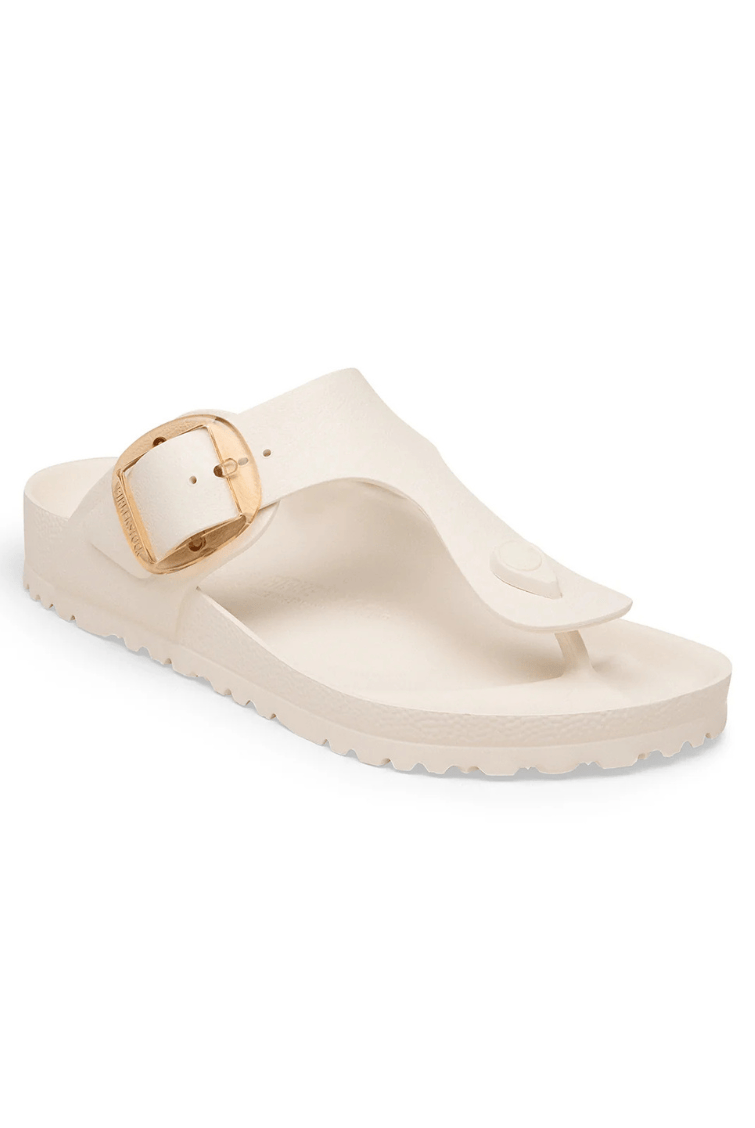 A single cream-colored thong sandal with a wide strap and a gold-tone buckle, shown from a side angle on a white background.