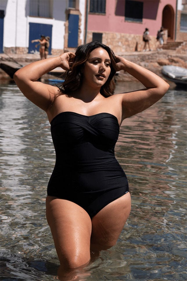 brunette model stands in knee deep water in spain in a plain black strapless one piece swimsuit