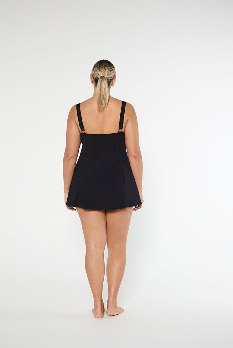 back of model wearing a black swim dress standing in front of a white wall