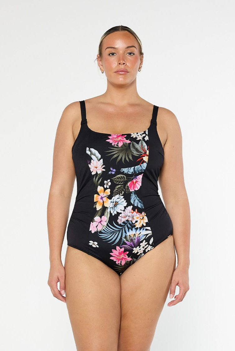 Blonde model shows the front of black floral one piece swimsuit with adjustable straps and supportive shelf bra