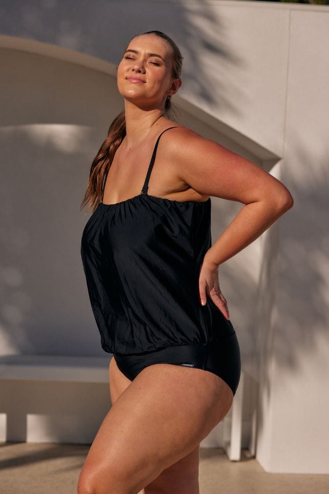 Blonde haired model stands in a black flouncy bandeau one piece. the swimsuit is worn strapless and has a loose top
