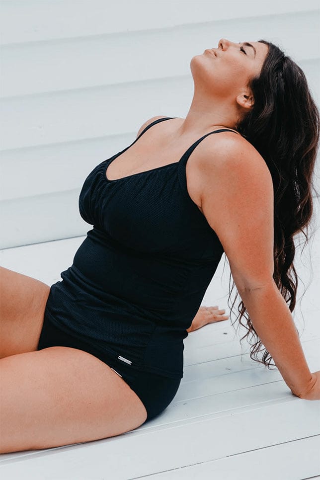 Model leans back in a sitting position againts a white background. The plus size model wears a black underwire tankini with honeycomb detail and underwire. She has styled this with a plain black bikini bottom