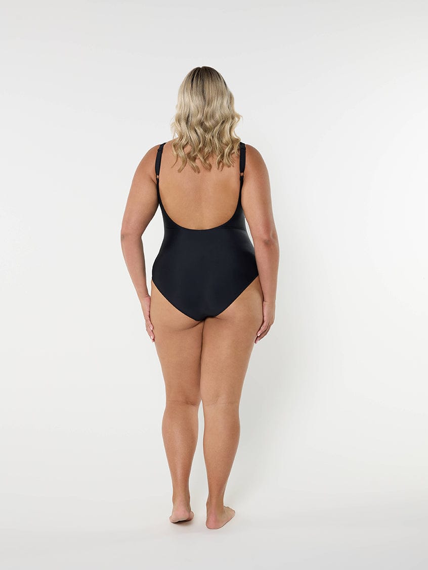 back of model standing in front of a white wall wearing a black one piece swimsuit