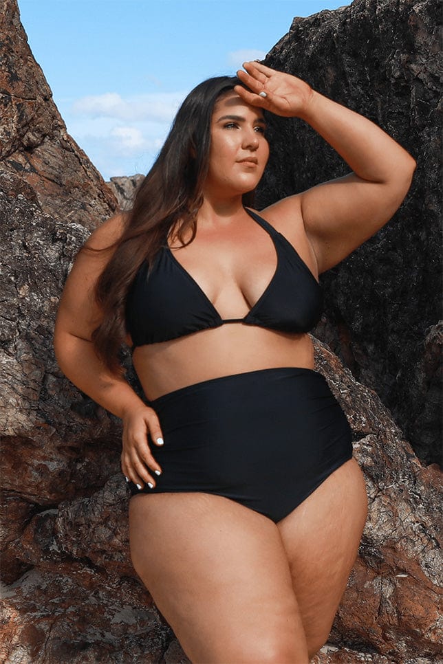 Model poses infront of rocks in a black triangle bikini top, the top ties at the neck and the back. She has paired this top with a black high waisted pant.