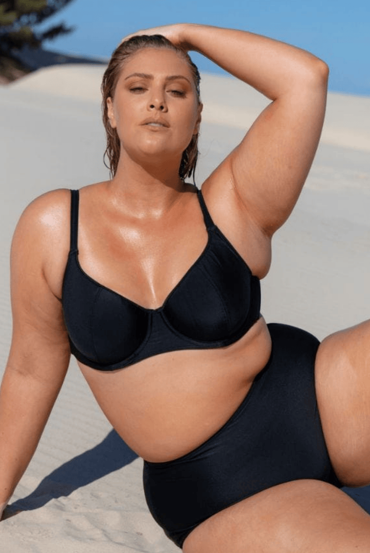 Model sits on the sand wearing a plain black underwire bikini top