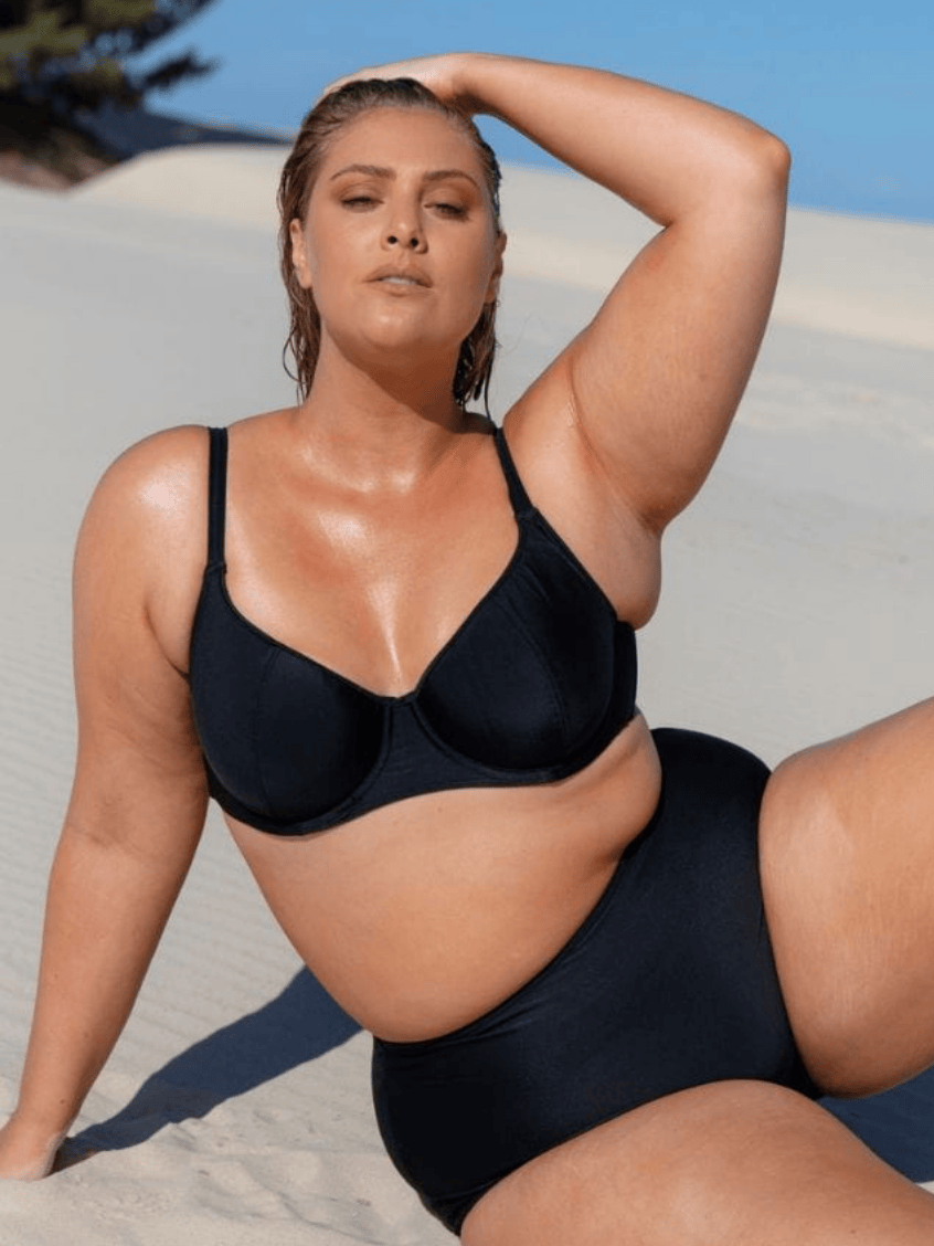 Model sits on the sand wearing a plain black underwire bikini top
