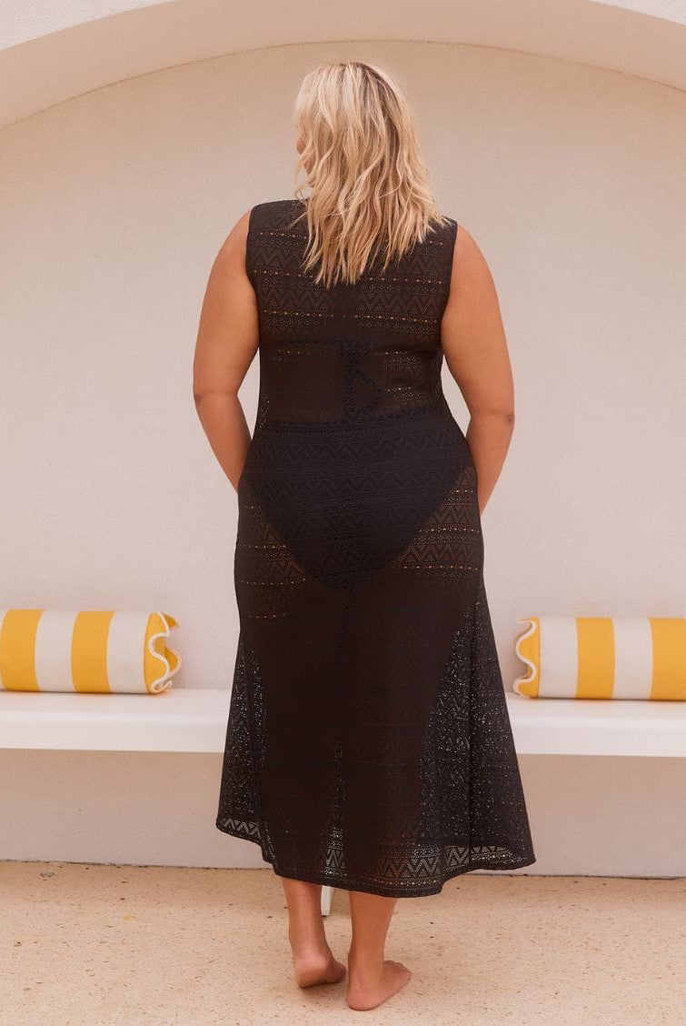 A woman stands with her back to the camera wearing a sleeveless black lace midi dress with a sheer back panel. She is standing in front of a white arched wall with yellow and white striped pillows on a bench.