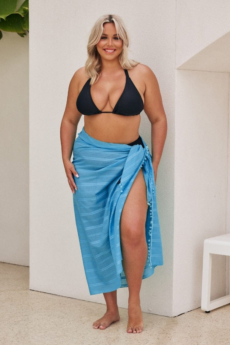 Woman wearing a black bikini top and blue sarong standing indoors.