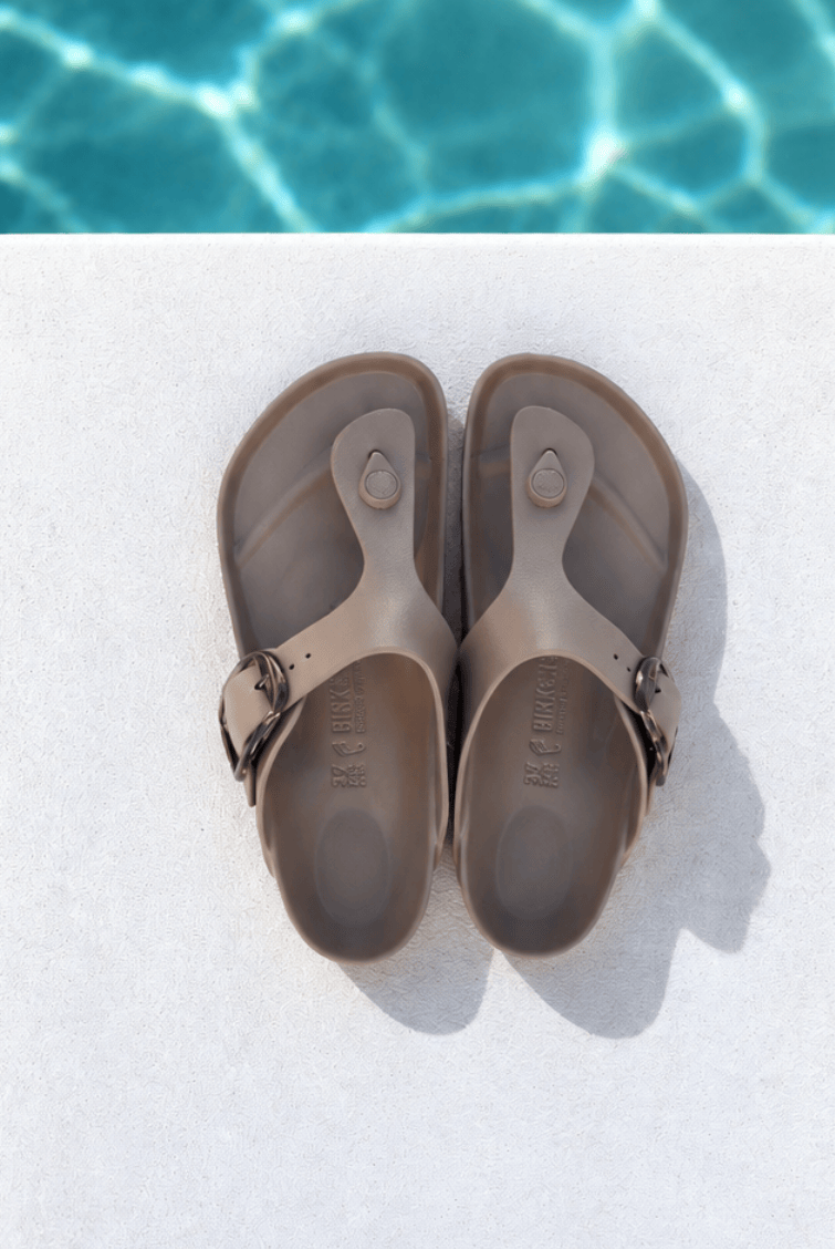 A pair of taupe-brown thong sandals with adjustable straps, laid flat on a white surface with a swimming pool background visible at the top.