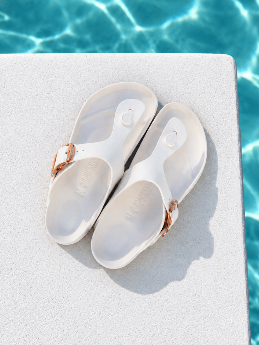 A pair of translucent white slide sandals with metallic rose gold buckle straps, resting on a white poolside deck with blue water in the background.