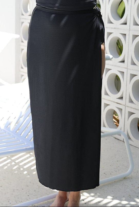 lady standing in front of a white walling wearing a long split swim skirt in black