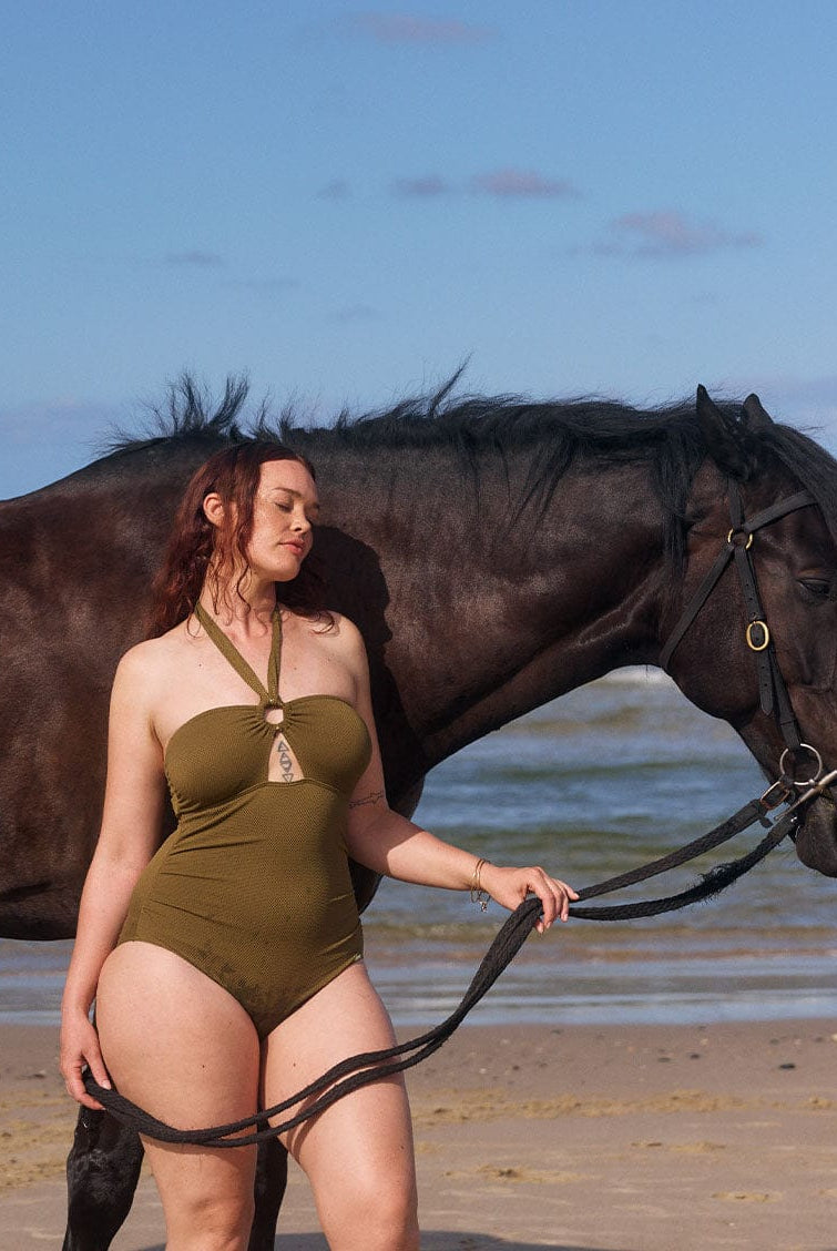 model stands next to a horse wearing figure flattering olive bandeau halter neck one piece with removable straps and power mesh front