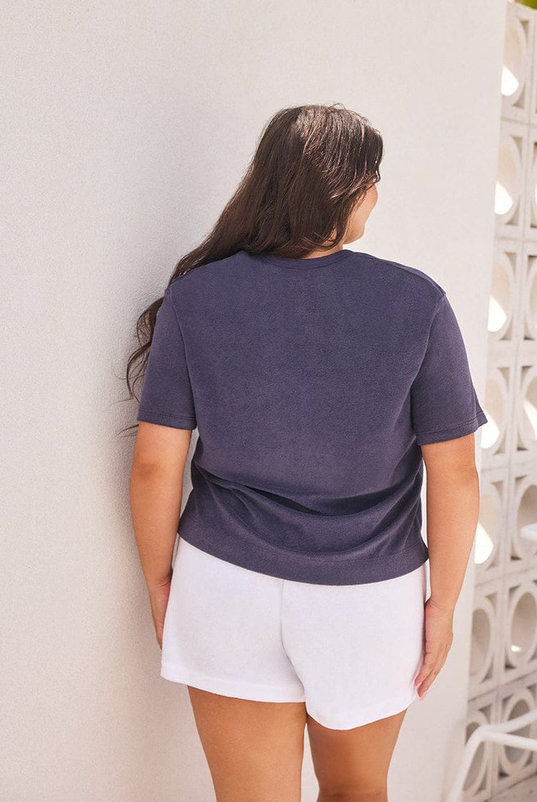 Person wearing a dark blue t-shirt and white shorts standing against a light-colored wall.