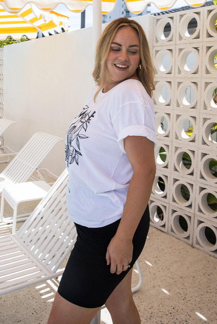 Woman wearing a white t-shirt and black shorts standing in front of a decorative wall with lounge chairs.