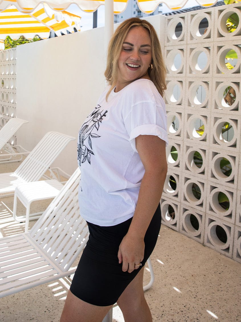 Woman wearing a white t-shirt and black shorts standing in front of a decorative wall with lounge chairs.