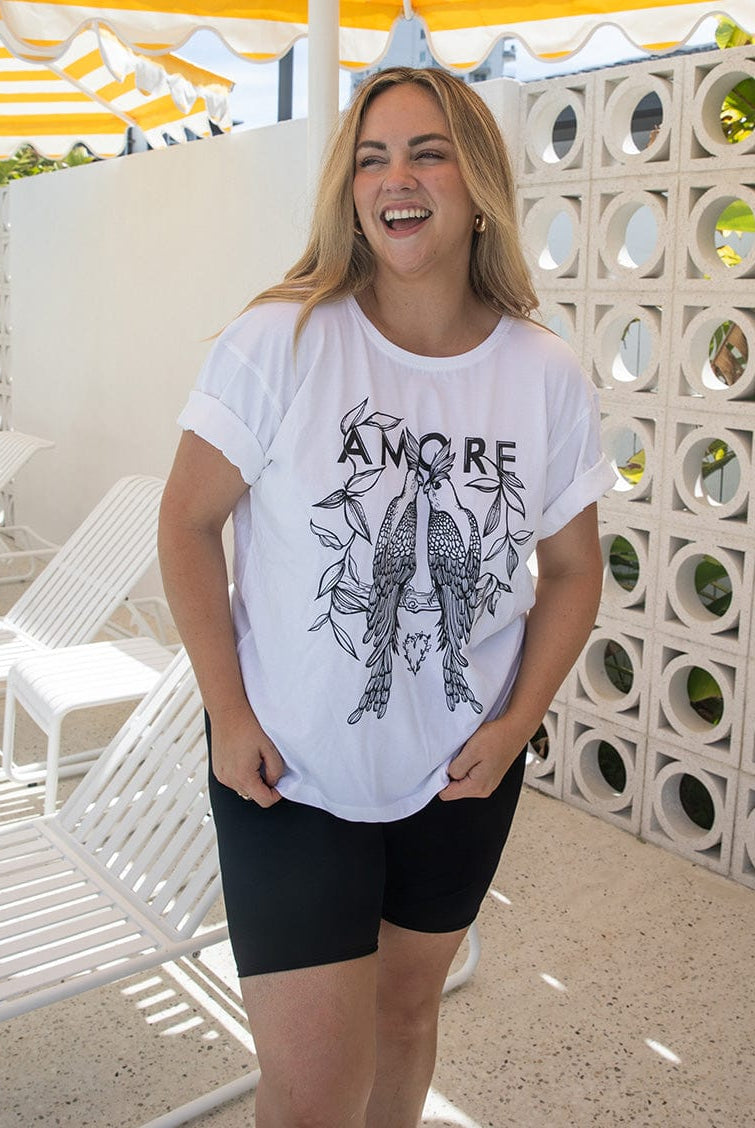 Woman wearing a white t-shirt with a graphic design and text, standing in a sunlit outdoor setting.