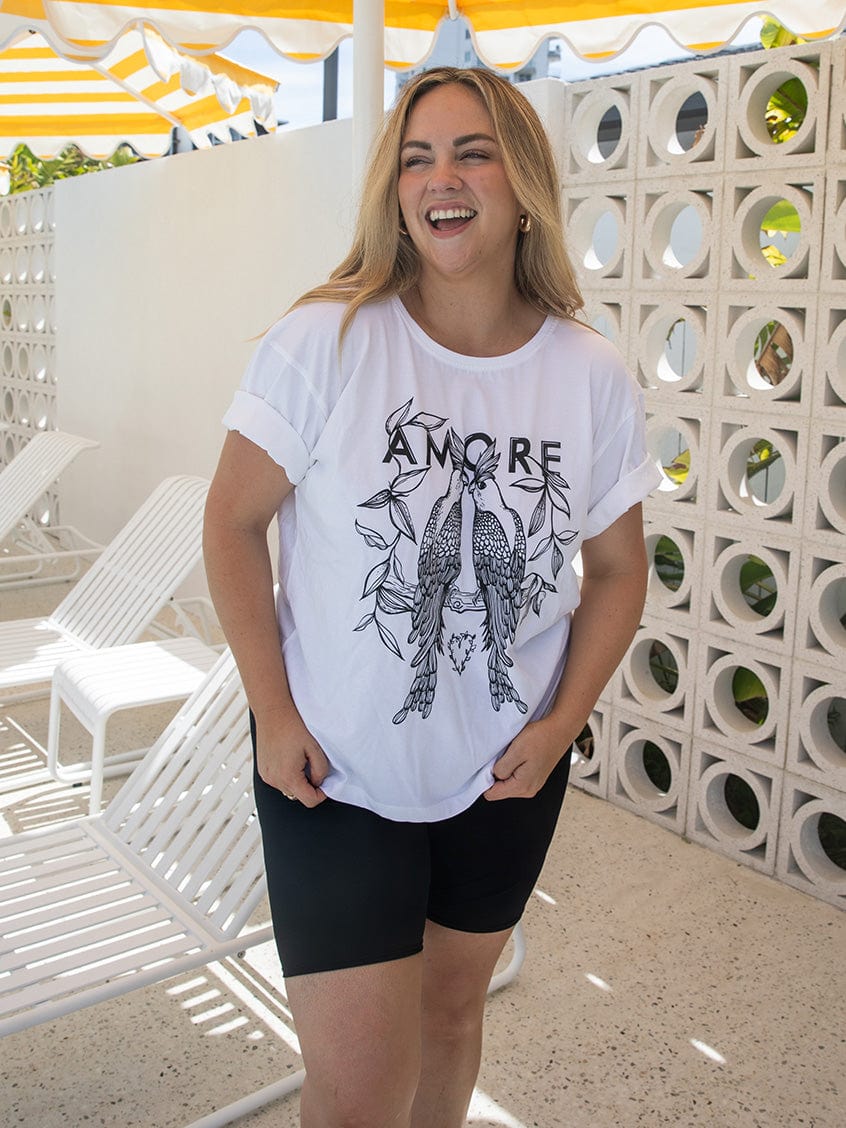 Woman wearing a white t-shirt with a graphic design and text, standing in a sunlit outdoor setting.