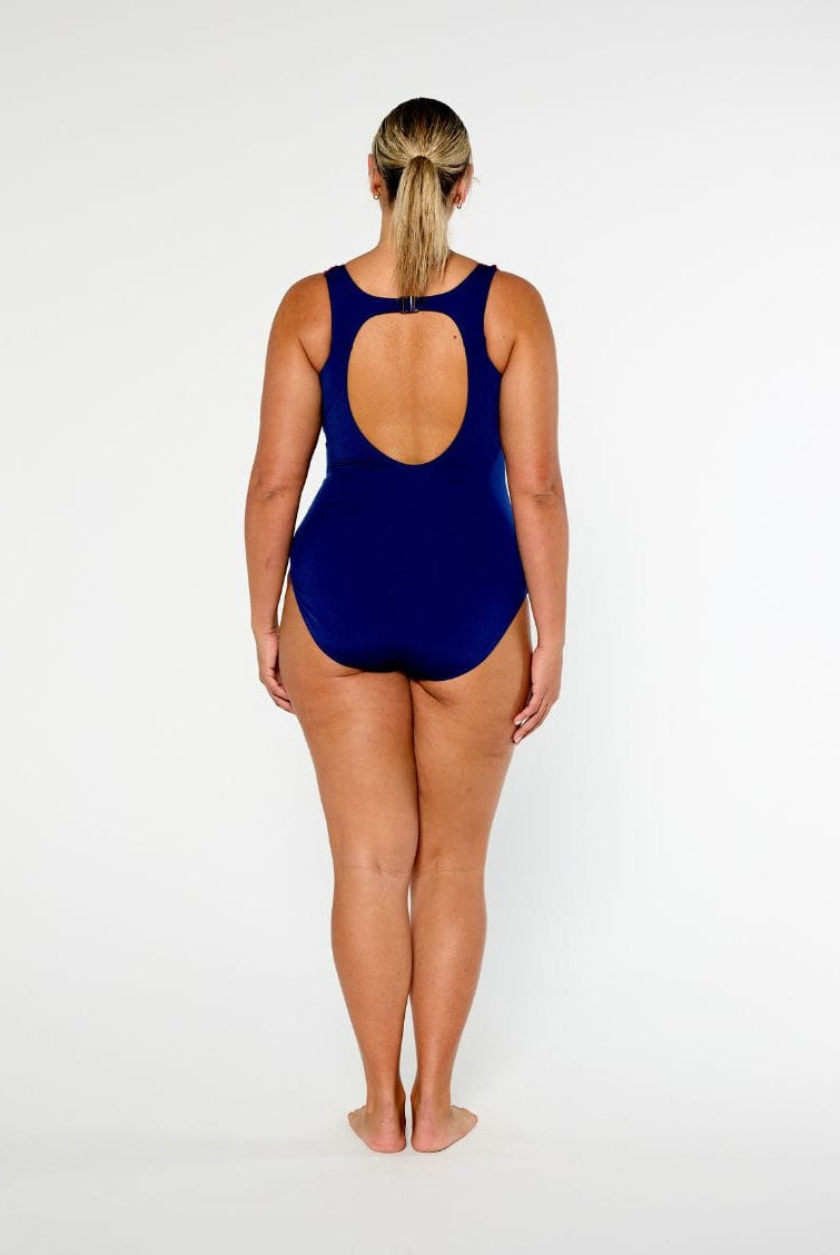 A woman standing with her back to the camera wearing a navy blue one-piece swimsuit with a large open back cutout, set against a plain white studio background.