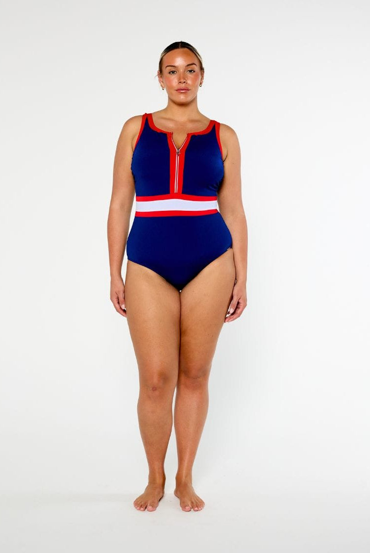 A woman modeling a one-piece swimsuit with a navy blue base and red and white striped accents on the neckline and waist, photographed against a plain light gray studio background.
