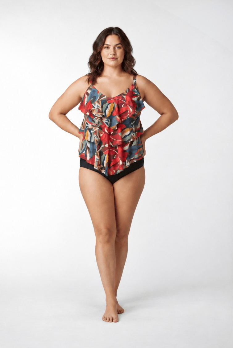 A woman modeling a ruffled tankini swimsuit with a vibrant multicolor abstract print and solid black bottoms, standing against a light gray studio background.