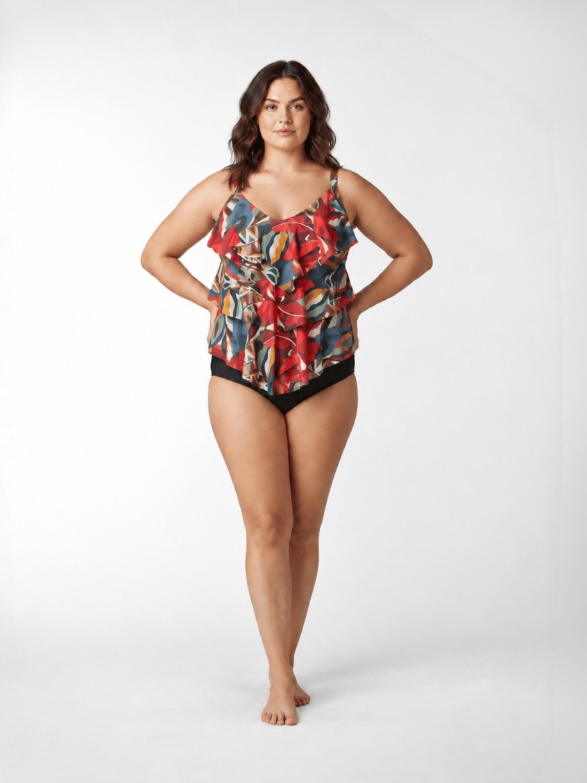 A woman modeling a ruffled tankini swimsuit with a vibrant multicolor abstract print and solid black bottoms, standing against a light gray studio background.