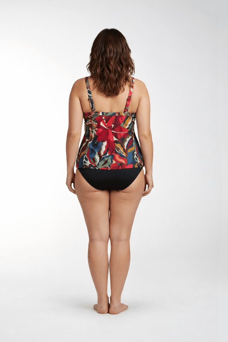 A woman is shown from behind wearing a one-piece swimsuit with a vibrant tropical floral print on the top and solid black bottoms, standing against a light gray studio background.