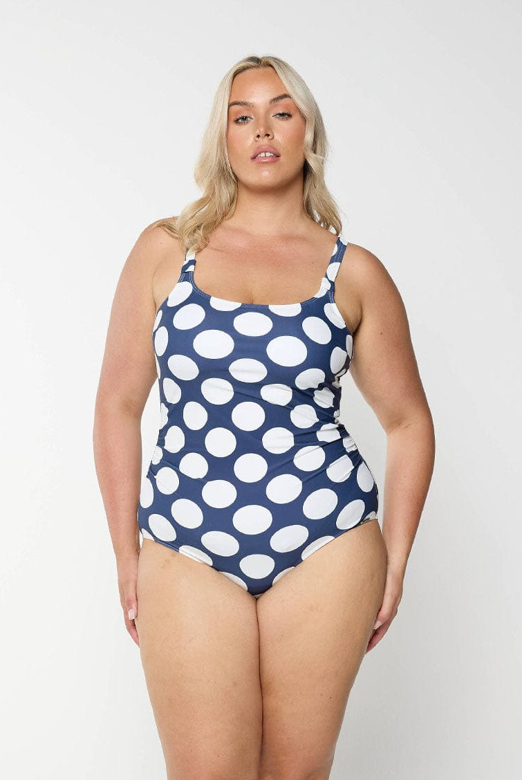 Blonde model shows the front of navy one piece with large white polkadots and a flattering scoop neckline