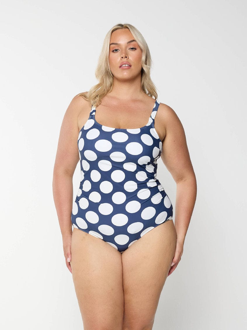 Blonde model shows the front of navy one piece with large white polkadots and a flattering scoop neckline