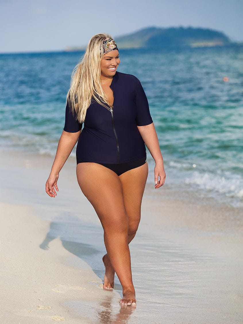 Woman in a navy swimsuit walking on a beach with ocean and sky in the background