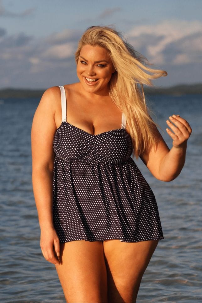 Blonde model smiles standing on the beach in a navy tankini top with white spots. The tankini swimsuit has removable straps, a fitted bust and loose middle.