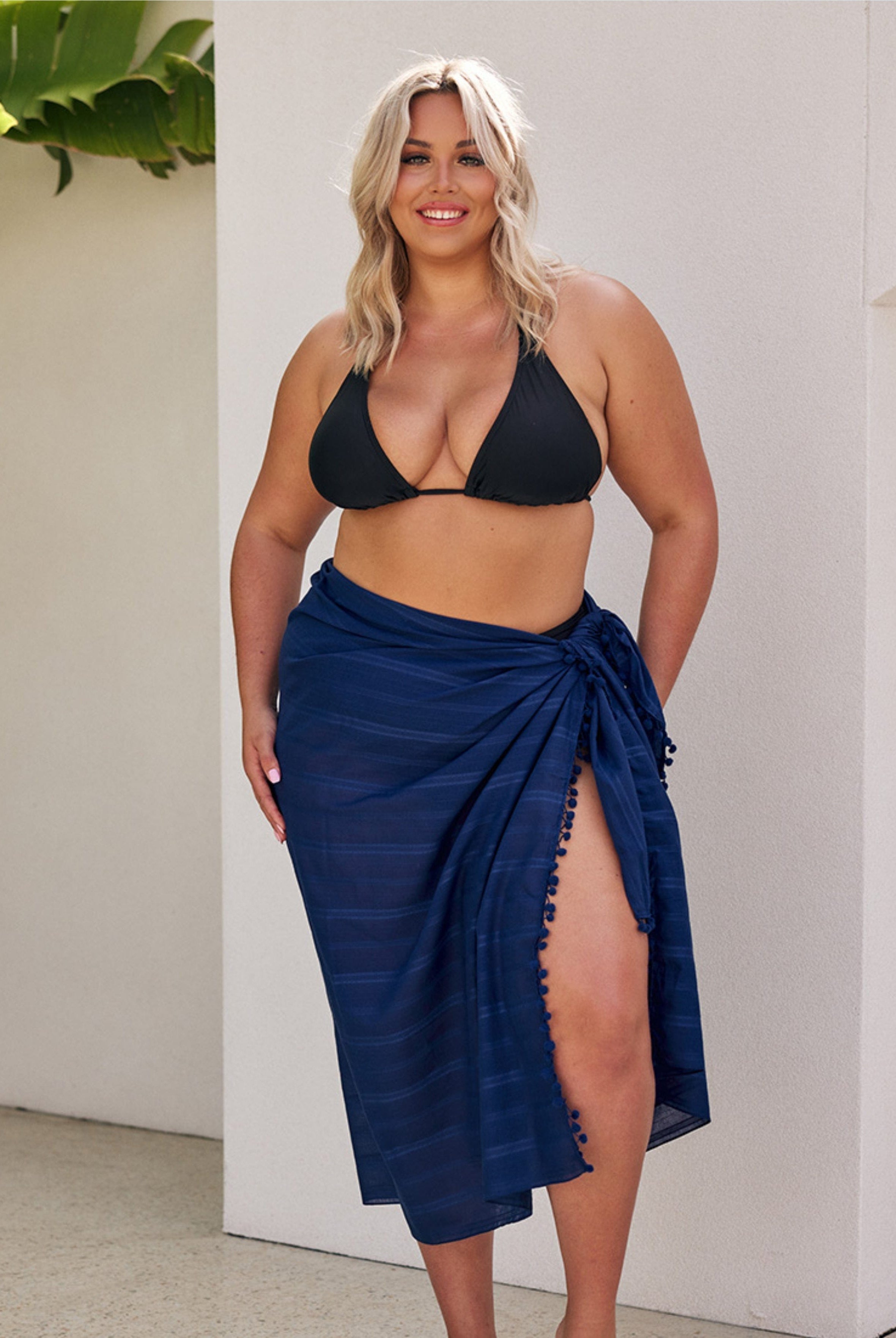 Woman wearing a black bikini top and navy blue sarong against a white wall.